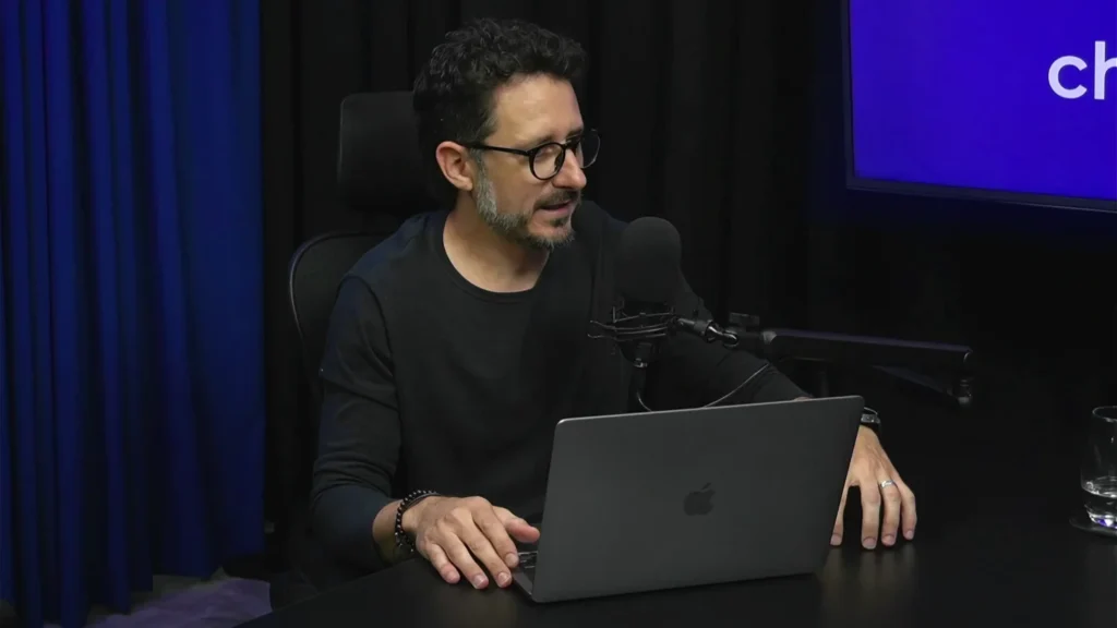 Piero Brustin, CBO do Chatspot, falando durante gravação de podcast com notebook Apple sobre a mesa, microfone profissional e tela ao fundo exibindo a marca Chatspot, em estúdio com cortina azul e iluminação escura